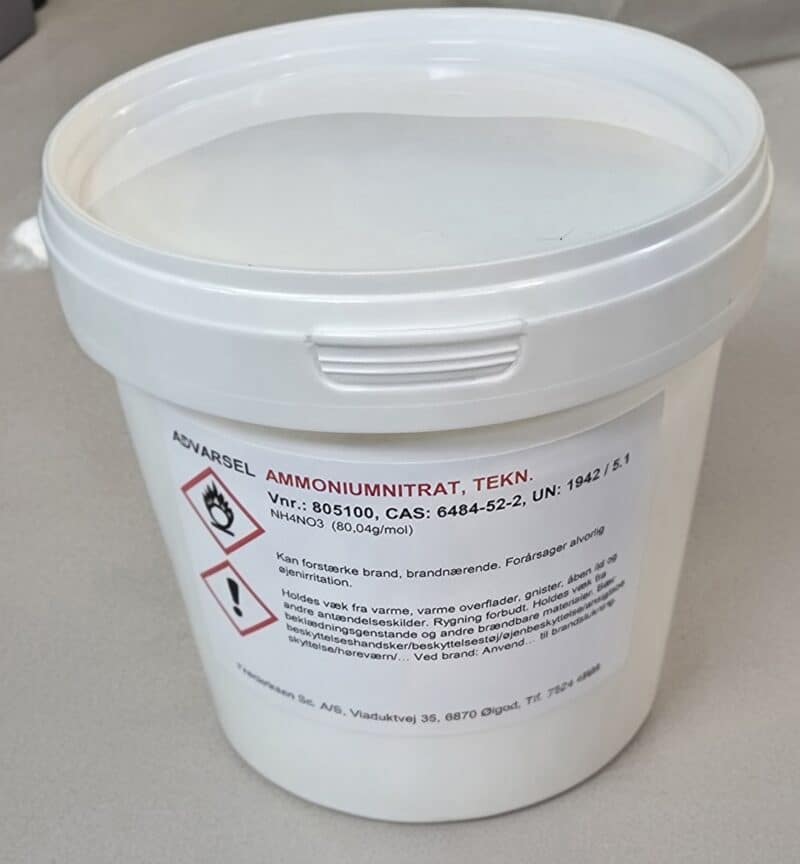 En hvid plastbeholder med Ammoniumnitrat 800 g med faresymboler og sikkerhedsoplysninger på etiketten, placeret på en grå overflade.