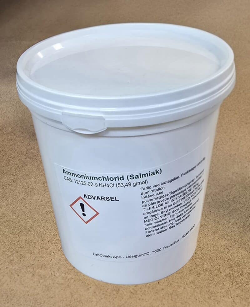 En hvid plastbeholder mærket "Ammoniumchlorid (800 g)" med advarselstekst og et faresymbol på en brun overflade.