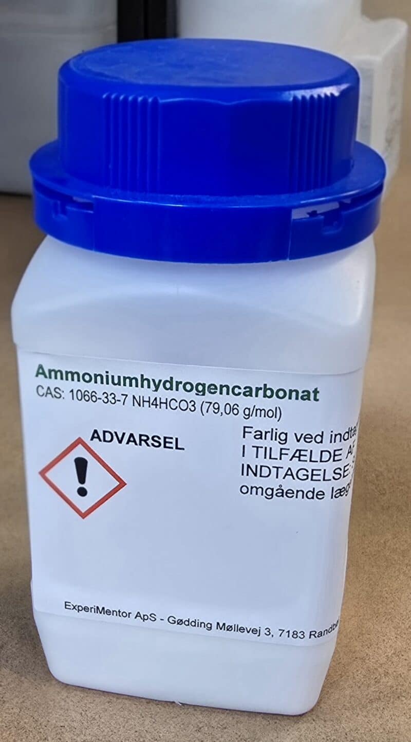 En hvid plastbeholder med blåt låg mærket Ammoniumhydrogencarbonat p.a. (500 g) og danske advarsler.