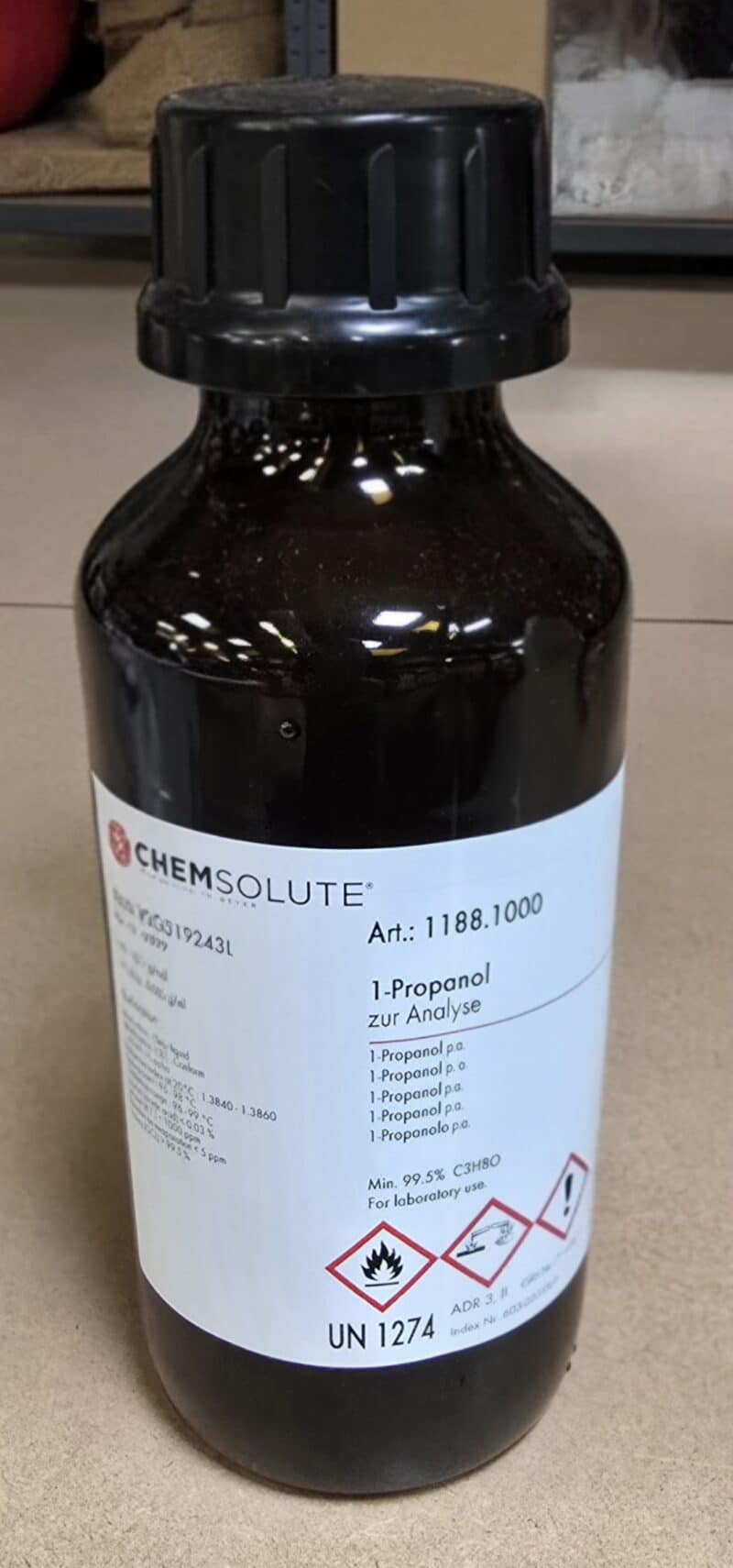 En mørk glasflaske mærket Propan-1-ol p.a. (1 liter) med faresymboler, stregkode og kemiske data på en hvid etiket.