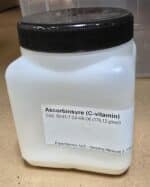 En hvid plastbeholder mærket "Ascorbinsyre, ren (200 g)" med kemiske oplysninger og virksomhedsoplysninger på en brun overflade.