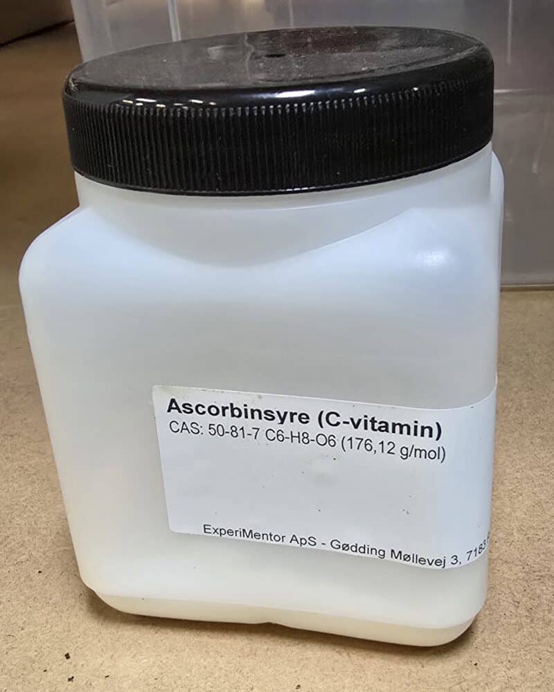 En hvid plastbeholder mærket "Ascorbinsyre, ren (200 g)" med kemiske oplysninger og virksomhedsoplysninger på en brun overflade.