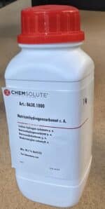 En hvid plastbeholder med 1 kg natriumhydrogencarbonat med rød hætte, mærket med kemikalie- og sikkerhedsoplysninger.