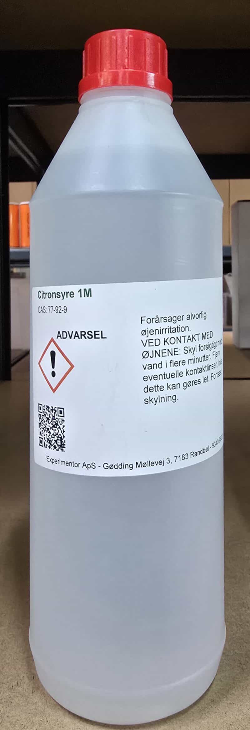 En hvid plastflaske med rød hætte mærket Citronsyre 1M (1 liter), advarsler og danske sikkerhedsanvisninger.