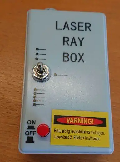 En grå Laserboks, elevsæt med vippekontakt, rød knap og gul advarselsmærkat hviler på en træoverflade.