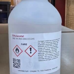 En hvid plastflaske med sort låg mærket Ethylacetat 1 L, 99,5%, med faresymboler og advarsler på dansk.