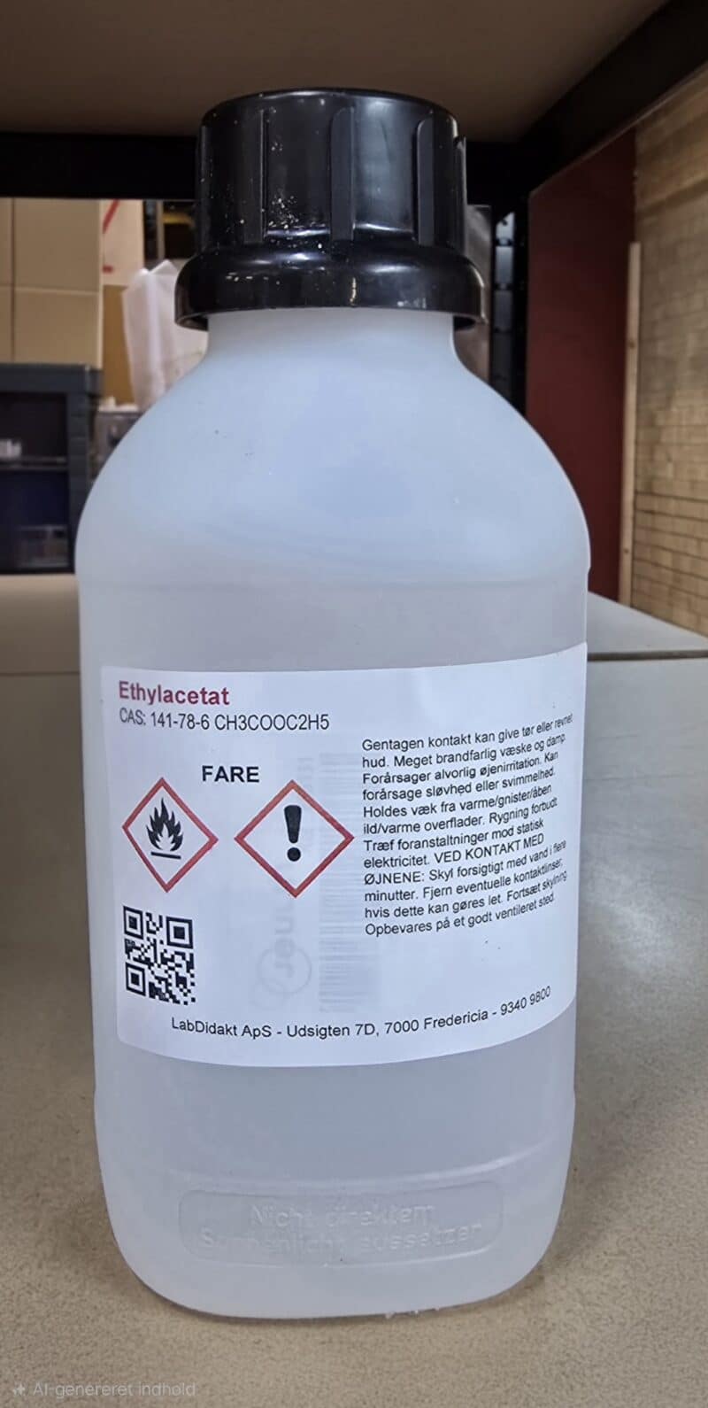 En hvid plastflaske med sort låg mærket Ethylacetat 1 L, 99,5%, med faresymboler og advarsler på dansk.