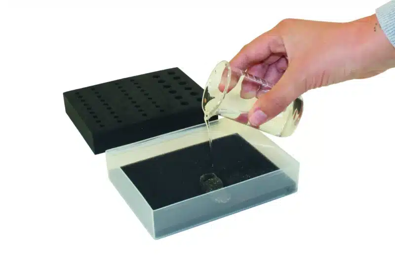 En hånd hælder væske i Kølet stativ til PCR-mikrorør - Stor model, ved siden af en sort holder med huller.