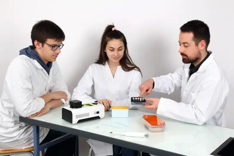 Tre laboranter diskuterer og undersøger reagensglas og et apparat, mens de bruger PCR Express Kit: Princip og implementering af PCR.