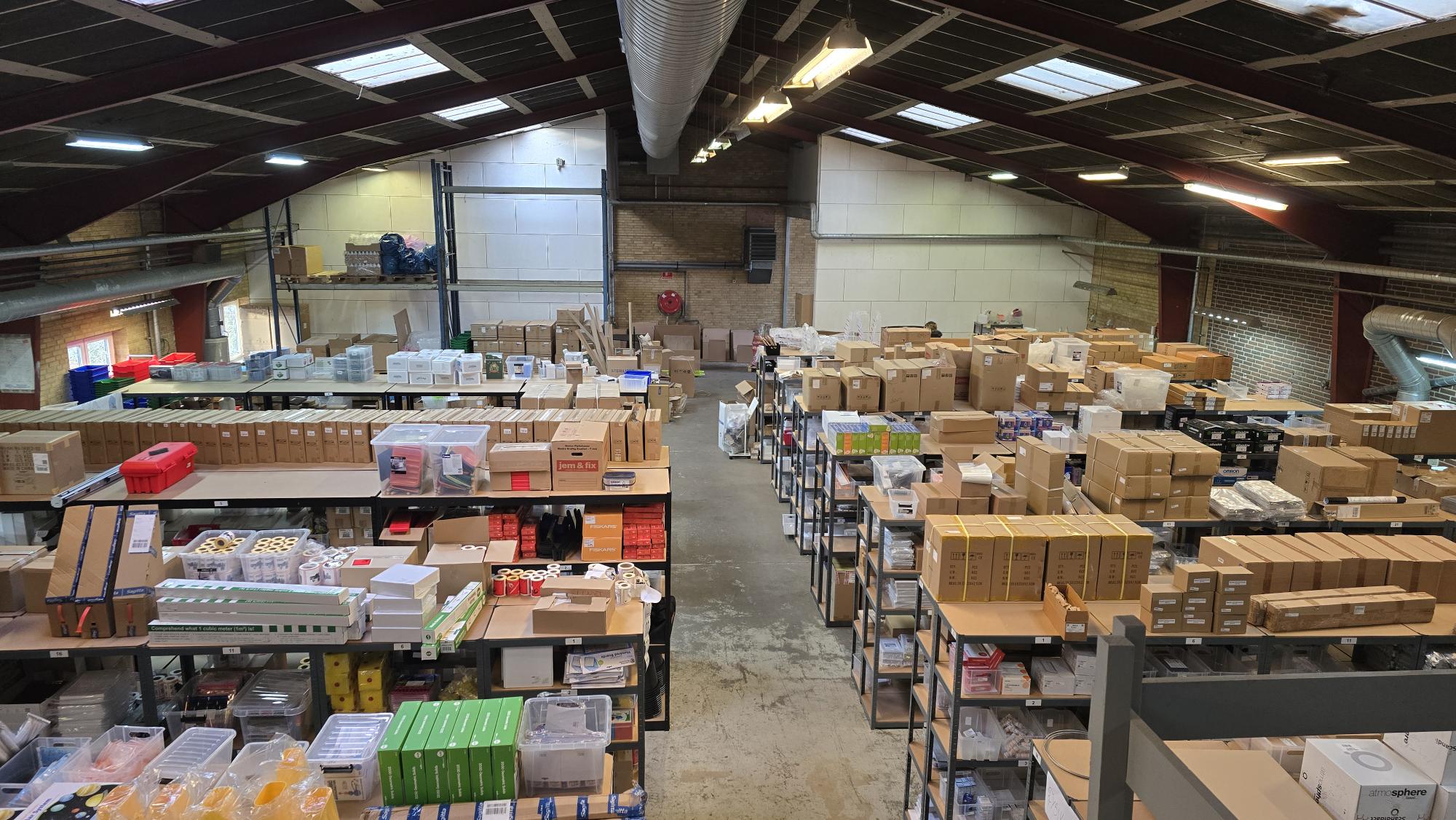 Et lagerinteriør med hylder fyldt med forskellige kasser, beholdere og pakker, set fra et højt perspektiv.