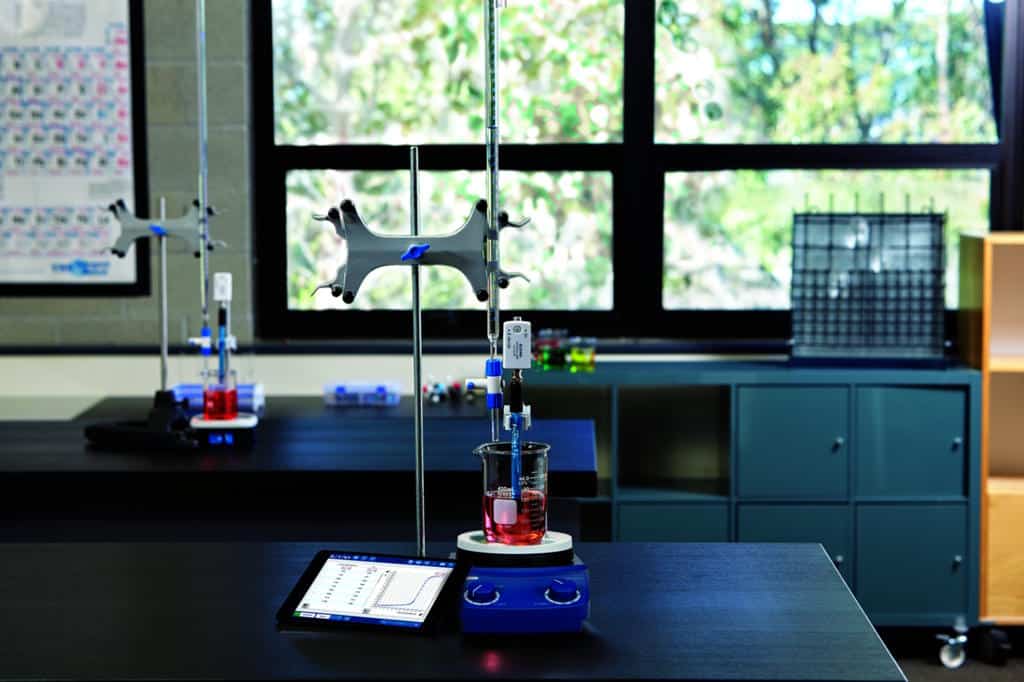 pasco-titrering - LabDidakt.dk Et bægerglas med rød væske står på en røreplade, med en burette over sig og en tablet i nærheden, i et lyst naturfagslokale.