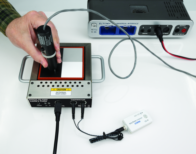 En hånd holder Leslies terning-sensoren (NY) over en metalplade, der er forbundet til laboratorieudstyr med kabler og en interface-enhed.