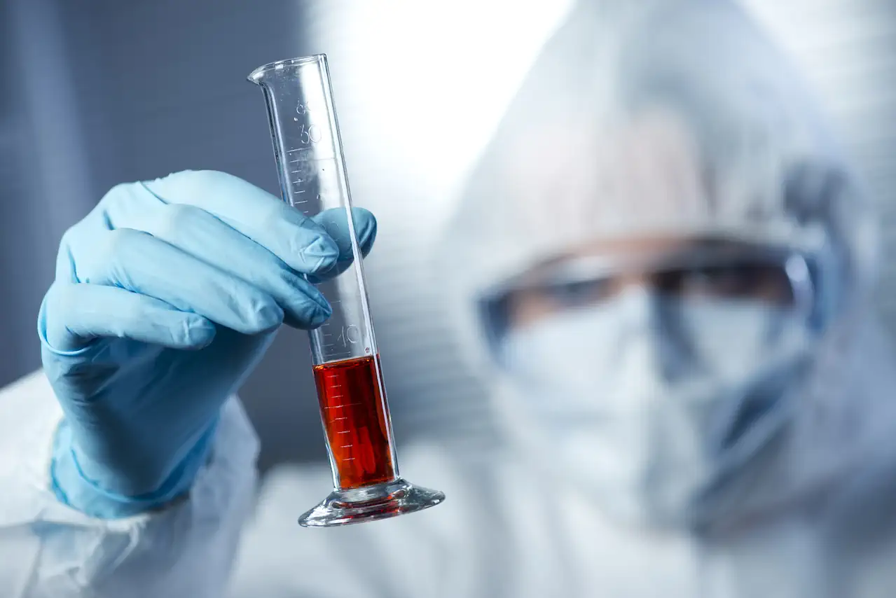 En person i beskyttelsesudstyr holder en målcylinder fyldt med rød væske i et laboratorium.