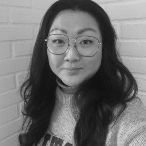 En kvinde med langt mørkt hår og briller bærer en sweater og står foran en hvid murstensvæg. Billedet er sort-hvidt.