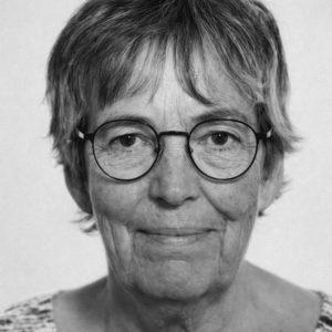 En ældre kvinde med kort gråt hår og runde briller kigger ind i kameraet og smiler lidt, vist i sort-hvid.